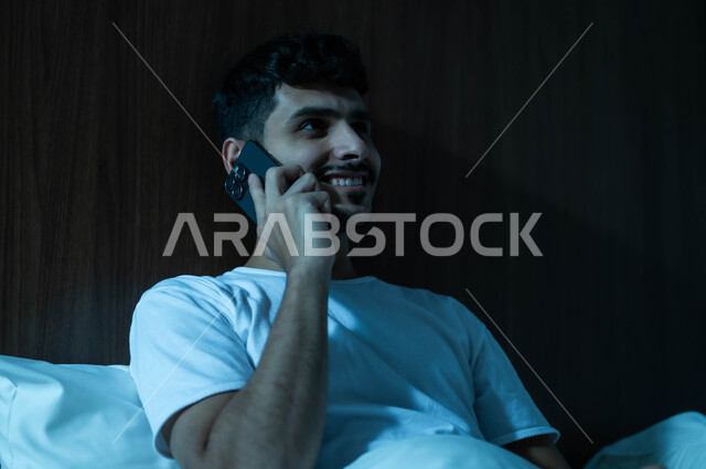 استخدام التقنيات والتكنولوجيا الحديثة المتطورة، التواصل مع الأهل والأصدقاء، صورة مقربة لرجل عربي خليجي سعودي مبتسم يجلس في سرير مريح يمسك بيده الهاتف المحمول يقوم بإجراء مكالمة هاتفية، الاستمتاع بالأجواء الهادئة