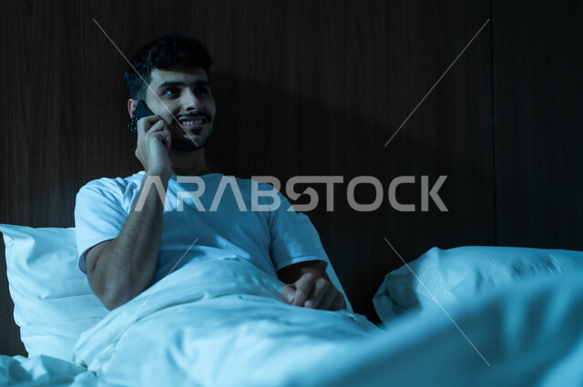 التواصل مع الأهل والأصدقاء، استخدام التكنولوجيا والتقنيات الحديثة المتطورة، رجل عربي خليجي سعودي مبتسم يجلس في سرير مريح يمسك بيده الهاتف المحمول يقوم بإجراء مكالمة هاتفية، الاستمتاع بالأجواء الهادئة