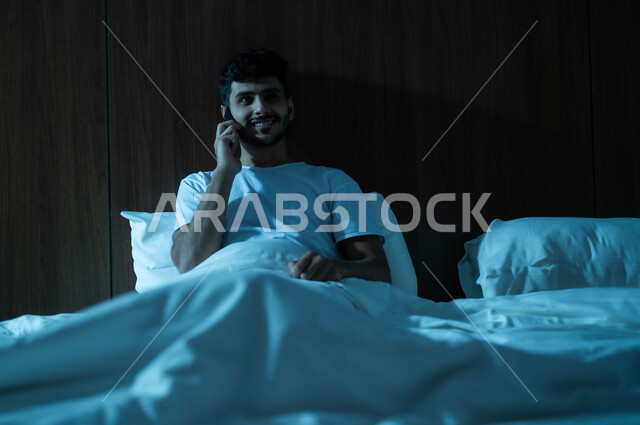 Using technology and advanced modern techniques, enjoying the quiet atmosphere, a smiling Saudi Arabian Gulf man sitting in a comfortable bed holding a mobile phone in his hand making a phone call, communicating with family and friend