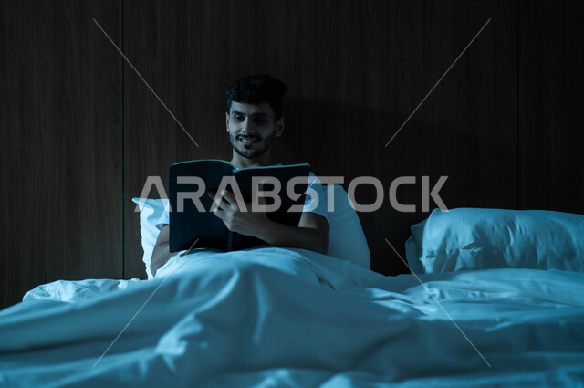 الاستمتاع بالأجواء الهادئة، قراءة القصص والروايات الممتعة، الشعور بالراحة والطمأنينة, رجل عربي خليجي سعودي مبتسم يجلس في سرير مريح يمسك بيده الكتاب، اتباع أسلوب حياة صحي