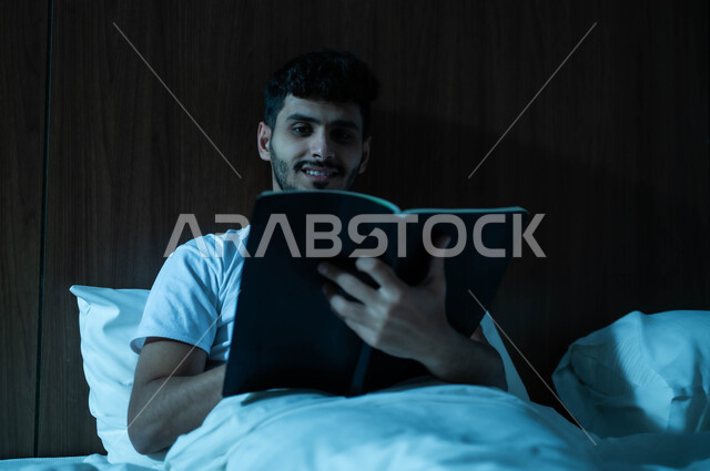 قراءة القصص والروايات الممتعة، الشعور بالراحة والطمأنينة, رجل عربي خليجي سعودي مبتسم يجلس في سرير مريح يمسك بيده الكتاب، الاسترخاء والاستمتاع بالأجواء الهادئة، اتباع أسلوب حياة صحي