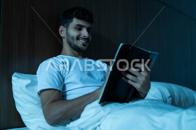 الاسترخاء والاستمتاع بالأجواء الهادئة، قراءة القصص والروايات الممتعة، الشعور بالراحة والطمأنينة, صورة مقربة لرجل عربي خليجي سعودي مبتسم يجلس في سرير مريح يمسك بيده الكتاب، اتباع أسلوب حياة صحي