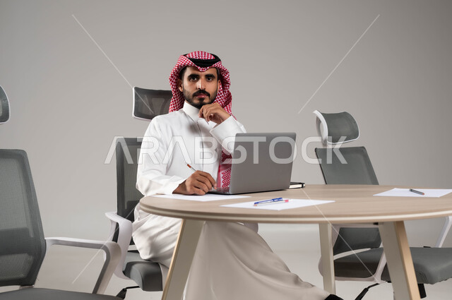 Using a laptop to accomplish the required tasks, a portrait of a Saudi Gulf Arab man wearing a shemagh and traditional dress, sitting at a meeting table, placing his hand under his head, looking at the camera with self-confidence gestures, taking important notes, gray background