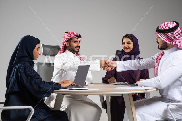 اتفاقيات وصفقات عمل جديدة، إيماءات التهنئة والنجاح في تحقيق اهداف الشركة, مفهوم روح الفريق والإنجاز الجماعي,  بورتريه مقرب لموظف عربي سعودي خليجي يقوم بمصافحة زميله, استخدام التكنولوجيا والتقنيات الحديثة، خلفية رمادية