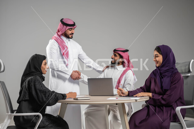 Using modern devices and technologies to complete office work, shaking hands with happy gestures and welcoming the new colleague, dialogues and discussions within the team, providing a suitable work environment, close-up portrait of Saudi Arabian Gulf employees in a meeting, success and achieving goals, gray background