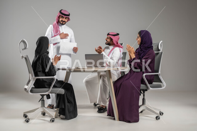 Gestures of joy and happiness, success in achieving the company’s goals and strategies, the concept of focus and accuracy in achievement, dialogues and discussions within the work team, a portrait of Saudi Gulf Arab employees applauding and congratulating their colleagues, workshops and training, gray background