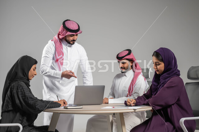 مفهوم التركيز والدقة في الإنجاز، الإشارة إلى اللابتوب بإيماءات الشرح وتوضيح المهام المطلوبة، الحوارات والنقاشات بين الزملاء، بورتريه لفريق عربي خليجي سعودي في اجتماع, ورشات عمل وتدريب، تدوين الملاحظات الهامة، خلفية رمادية