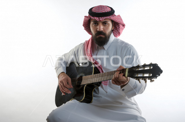 بورتريه شاب سعودي عربي خليجي يعزف على  الجيتار( آلة وترية) ، شاب باللباس السعودية التقليديه بخلفية بيضاء ، آلة موسيقية ، مجال صوتي و فني