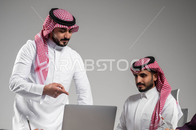 الإشارة إلى الحاسب المحمول بإيماءات الشرح والتوضيح, دراسة ومناقشة المشاريع الجديدة, مهن ووظائف سعودية، بورتريه مقرب لرجل عربي خليجي سعودي يرتدي الشماغ والثوب التقليدي يقوم بتدريب زميله، إستخدام التقنيات والتكنولوجيا الحديثة، خلفية رمادية