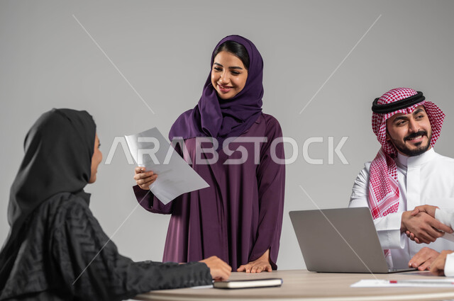 Saudi professions and jobs, providing a comfortable work environment, dialogues and agreements within the team, portrait of a smiling Saudi Arabian Gulf employee holding papers in her hand discussing with her colleagues with gestures of pleasure, using modern techniques and technology, gray backgroun