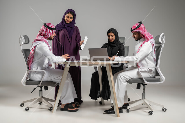 Business and company development, business meeting and project completion, focus and careful work, dialogues and discussions within the team, discussion of plans and explanation of business tasks, portrait of Saudi Gulf Arab co-workers in a meeting, wearing traditional Saudi dress, gray background
