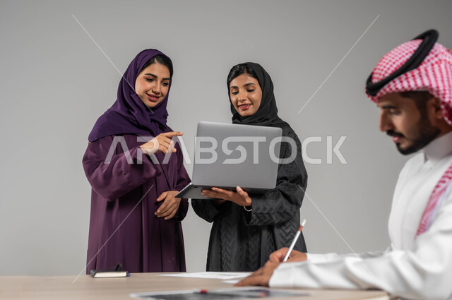 توجيه الجهود لتحقيق أهداف الشركة, الإشارة الى الحاسوب المحمول بهدف توضيح العمليات, مفهوم الشراكة والتعاون, بورتريه مقرب لزملاء عرب سعوديين خليجيين في اجتماع, ارتداء اللباس السعودي التقليدي, خلفية رمادية