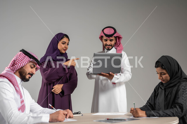 التفاهم والتعاون بين الزملاء لتحقيق الأهداف, استخدام الاجهزة الحديثة في تطوير الأعمال, بورتريه مقرب لموظفين عرب سعوديين خليجيين في اجتماع, ارتداء اللباس السعودي التقليدي, الاشارة الى اللابتوب بهدف توضيح العمليات والانجازات, خلفية رمادية