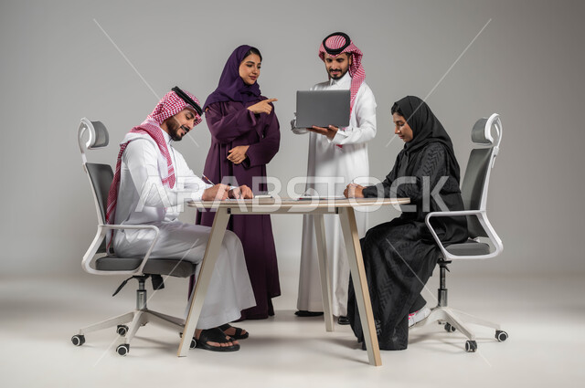 الاشارة الى الحاسوب المحمول بهدف توضيح العمليات, توفير بيئة عمل مناسبة, مفهوم الشراكة والعمل الجماعي, بورتيه لموظفين عرب سعوديين خليجيين في اجتماع, ارتداء اللباس السعودي التقليدي, استخدام الاجهزة والتقنيات الحديثة في تطوير الاعمال, خلفية رمادية