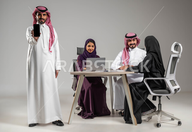 دمج التكنولوجيا الحديثة بالعمل، عرض شاشة الموبايل، إبداء الرأي ومشاركة الأفكار، الحوارات والنقاشات ضمن الفريق، ضمان فهم واضح للاعمال، التركيز والعمل الجاد، بورتريه لموظف عربي خليجي سعودي في اجتماع يحمل بيده الهاتف المحمول، صورة لكامل الجسم، خلفية رمادية