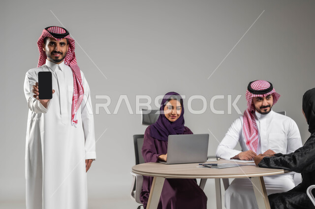 Gestures of happiness and pleasure, displaying the mobile screen, integrating modern technology into work, expressing opinions and sharing ideas, dialogues and discussions within the team, ensuring a clear understanding of the work, portrait of a Saudi Gulf Arab employee in a meeting holding a mobile phone in his hand, Saudi professions and jobs, gray background.