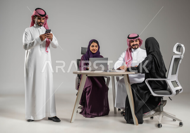 مهن ووظائف سعودية، الحوارات والنقاشات ضمن الفريق، إنجاز العمل عبر الموبايل، مفهوم الشراكة والتعاون، إستخدام التكنولوجيا الحديثة، الاحترافية والتفاني في العمل، بورتريه لموظفة عربية سعودية خليجية مقوم باجتماع مع زملائها، صورة لكامل الجسم، خلفية رمادية