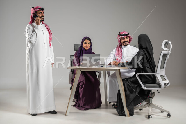 إستخدام التكنولوجيا الحديثة، إجراء مكالمات عمل، الاحترافية والتفاني في العمل، بورتريه لموظفة عربية سعودية خليجية مقوم باجتماع مع زملائها، مهن ووظائف سعودية، الحوارات والنقاشات ضمن الفريق، توفير بيئة عمل مناسبة، صورة لكامل الجسم، خلفية رمادية