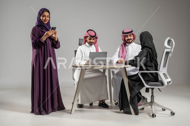 Dialogues and discussions within the team, completing work via mobile phone, the concept of partnership and cooperation, the use of modern technology, professionalism and dedication to work, a portrait of a Saudi Arabian Gulf employee having a meeting with her colleagues, Saudi professions and jobs, a full-body portrait, gray background.