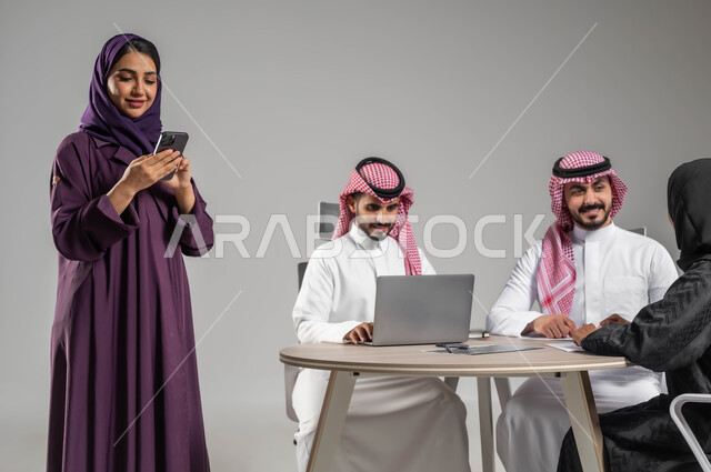إنجاز العمل عبر الموبايل، مفهوم الشراكة والتعاون، إستخدام التكنولوجيا الحديثة، الاحترافية والتفاني في العمل، بورتريه لموظفة عربية سعودية خليجية مقوم باجتماع مع زملائها، مهن ووظائف سعودية، الحوارات والنقاشات ضمن الفريق، توفير بيئة عمل مناسبة، خلفية رمادية