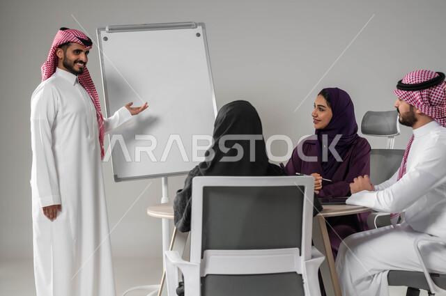 التفاني في الإنجاز، شرح مهام الأعمال، مفهوم التعاون وروح الفريق، استخدام السبورة لتوضيح العمليات والاجراءات المتبعة في الشركة، الاشارة إلى اللوح بهدف التوضيح، مناقشة خطط العمل، ضمان فهم واضح للاعمال، بورتريه لفريق عربي خليجي سعودي في اجتماع، خلفية رمادية