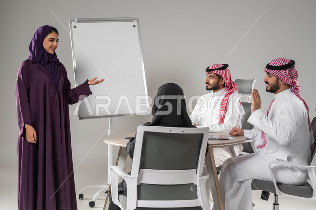 رفع اليد بهدف طلب الاذن لابداء الرأي، استعمال السبورة لتوضيح العمليات والاجراءات المتبعة في الشركة، الاشارة إلى اللوح بهدف التوضيح، مناقشة خطط العمل، ضمان فهم واضح للاعمال، الاحترافية والتفاني في الإنجاز، بورتريه لفريق عربي خليجي سعودي في اجتماع، خلفية رمادية