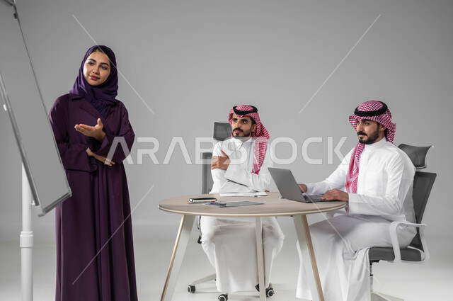Using the blackboard to explain the processes and procedures followed in the company, referring to the board for clarification, discussing work plans, ensuring a comprehensive understanding of the business, using the laptop to take notes, professionalism and dedication to achievement, a portrait of a Saudi Gulf Arab team in a meeting, gray background