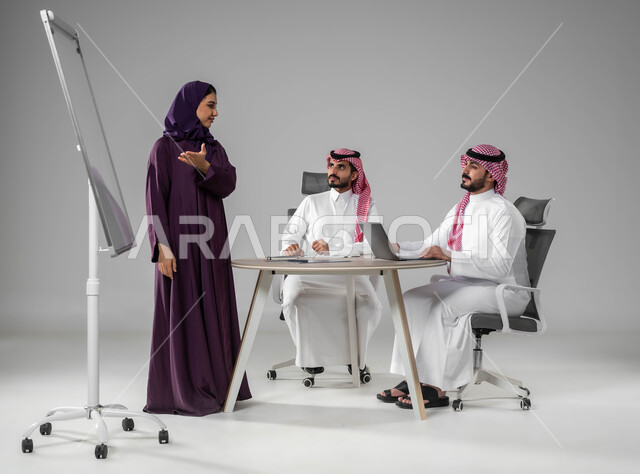 Ensuring a clear and comprehensive understanding of the business, using the blackboard to clarify the processes and procedures followed in the company, using the laptop to take notes, dedication to achievement, explaining business tasks, a portrait of a Saudi Gulf Arab team in a meeting, discussing work plans, a full-body portrait, gray background