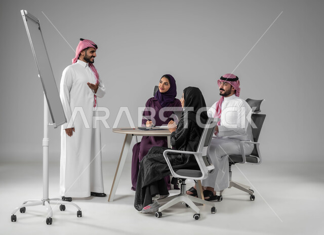 استخدام السبورة لتوضيح العمليات والاجراءات المتبعة في الشركة، استعمال اللابتوب في تدوين الملاحظات، ضمان فهم واضح وشامل للاعمال، التفاني في الإنجاز، شرح مهام الاعمال، بورتريه لفريق عربي خليجي سعودي في اجتماع، مناقشة خطط العمل، صورة لكامل الجسم، خلفية رمادية