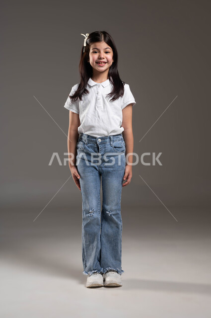التقاط صور شخصية للوثائق المدرسية, الإهتمام بالمظهر الخارجي للأطفال الصغار، بورتريه لطفلة عربيه خليجيه سعودية تنظر إلى الكاميرا مبتسمة ترتدي زي كاجوا، ارتداء الملابس الجديده في الأعياد والمناسبات، خلفيه رمادية، ايماءات تدل على السعادة، صورة الجسم بالطول الكامل