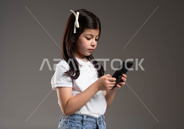 مشاهدة البرامج المفضلة عبر الانترنت، ايماءات الاندماج والتركيز، بورتريه مقرب لفتاه عربيه سعوديه خليجيه ترتدي الزي الكاجوال تقوم بتصفح مواقع التواصل الاجتماعي باستخدام الجوال، خلفية رمادية