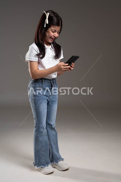 The role of modern technology in providing greater comfort for individuals, browsing the Internet and completing tasks remotely, portrait of a smiling Saudi Arabian Gulf girl wearing a casual outfit using a mobile phone, full-length body photo, gray background
