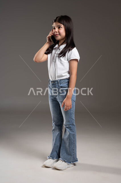 الدردشة والتواصل مع العائلة والأصدقاء ، استخدام التقنية والأجهزة الحديثة، بورتريه لفتاه عربيه سعوديه  خليجيه مبتسمة ترتدي الزي الكاجوال تتحدث على الهاتف المحمول، صورة الجسم بالطول الكامل، خلفية رمادية