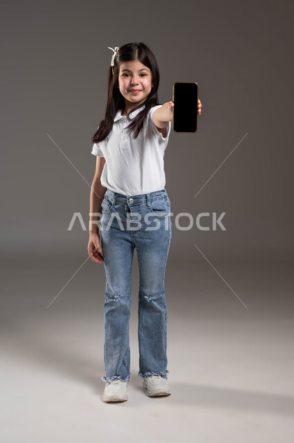 Using modern technology and techniques, e-marketing and displaying mobile services and applications, a portrait of a smiling Saudi Arabian Gulf girl wearing a casual outfit, holding a mobile phone in her hand, with a blank black screen, a full-body portrait, a gray background.