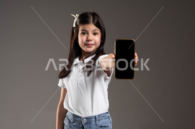 E-marketing and display of mobile services and applications, use of technology and modern technology, close-up portrait of a smiling Saudi Arabian Gulf girl wearing a casual outfit holding a mobile phone in her hand with a blank black screen, gray background