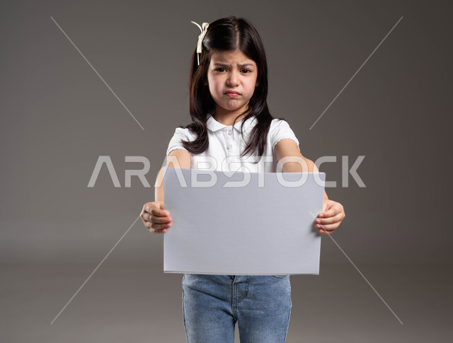 موك اب مربع صغير، الترويج للخدمات والمنتجات، بورتريه لفتاة عربية خليجية سعودية ترتدي الزي الكاجوال تمسك بيدها لوحة بيضاء فارغة وتنظر الى الكاميرا بتعابير الاستياء وعدم الرضا، خلفية رمادية