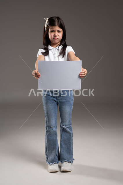 الترويج للخدمات والمنتجات، موك اب مربع صغير، بورتريه لفتاة عربية خليجية سعودية ترتدي الزي الكاجوال تمسك بيدها لوحة بيضاء فارغة وتنظر الى الكاميرا بتعابير الغضب وعدم الرضا، صورة لكامل الجسم، خلفية رمادية
