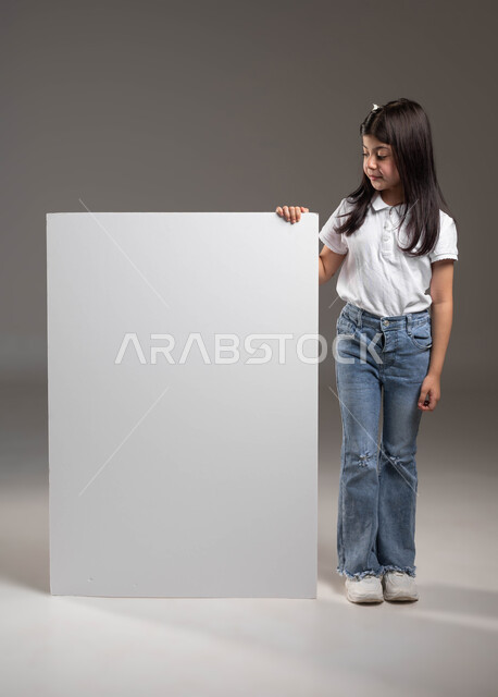 الترويج للخدمات والمنتجات، مفهوم الاعلان والتسويق، بورتريه لفتاة عربية خليجية سعودية ترتدي الزي الكاجوال تنظر إلى لوحة اعلانات بيضاء فارغة، موك اب ابيض مستطيل،  صورة الجسم بالطول الكامل، خلفية رمادية