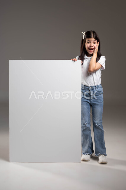 ايماءات التعجب والمفاجأة، مفهوم الاعلان والتسويق، الترويج للخدمات والمنتجات من خلال لوحة بيضاء فارغة، بورتريه لفتاة عربية خليجية سعودية ترتدي الزي الكاجوال تضع يدها على موك اب مستطيل كبير وتنظر الى الكاميرا بتعابير الدهشة، صورة لكامل الجسم، خلفية رمادية