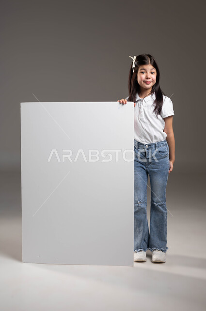 موك اب مستطيل كبير، إعلانات ودعايات وعروض تسويقية، إستخدام لوحات توضيحيه، بورتريه لفتاة عربية خليجية سعودية مبتسمة ترتدي الزي الكاجوال تضع يدها على لوحة بيضاء فارغة،  صورة الجسم بالكامل، خلفية رمادية