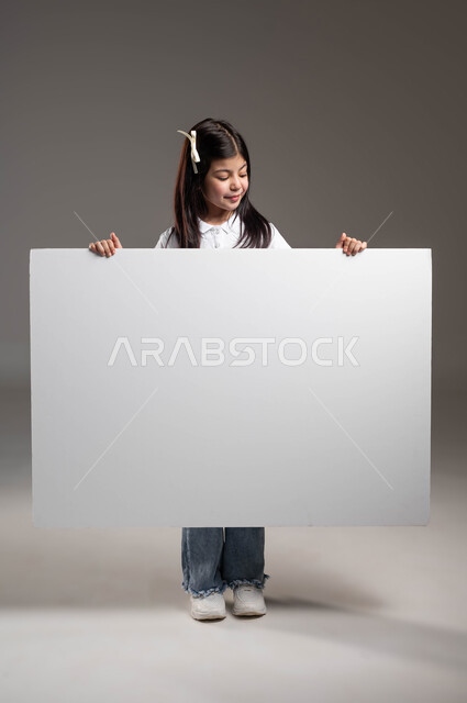 إستخدام لوحات توضيحيه، موك اب أبيض مستطيل ، اعلانات ودعايات وعروض تسويقية، بورتريه لفتاة عربية خليجية سعودية ترتدي الزي الكاجوال  تمسك بيدها لوحة بيضاء فارغة وتنظر اليها بتعابير السرور، خلفية رمادية