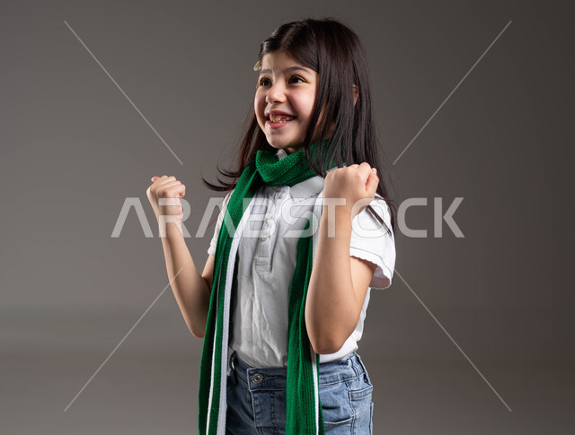 رفع اليد بكف مقبوض للأعلى بتعابير الانتصار، تشجيع الفريق السعودي لكرة القدم ، أنشطة رياضية ترفيهية للأطفال, بورتريه لفتاة عربية خليجية سعودية ترتدي الوشاح التشجيعي لفريقها المفضل وتحمل بيدها كرة قدم, إيماءات تدل على السرور والحماس لتحقيق الفوز و الانتصار، خلفية رمادية
