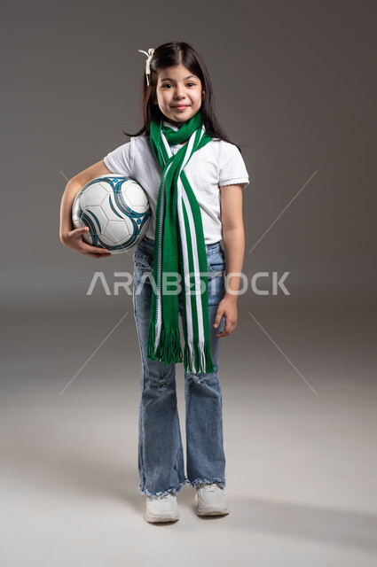 Wearing cheerleading scarves for the favorite team, a portrait of a smiling Saudi Gulf Arab girl wearing a casual outfit, holding a soccer ball in her hand, the passion for following sports clubs since childhood, looking at the camera with gestures of pleasure and enthusiasm to achieve victory and triumph, full-length body portrait, gray background