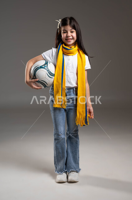 Wearing promotional scarves for the favorite team, a portrait of a smiling Saudi Gulf Arab girl wearing a casual outfit, holding a soccer ball in her hand, passion for following sports clubs since childhood, looking at the camera with gestures of pleasure and enthusiasm to achieve victory and triumph, gray background