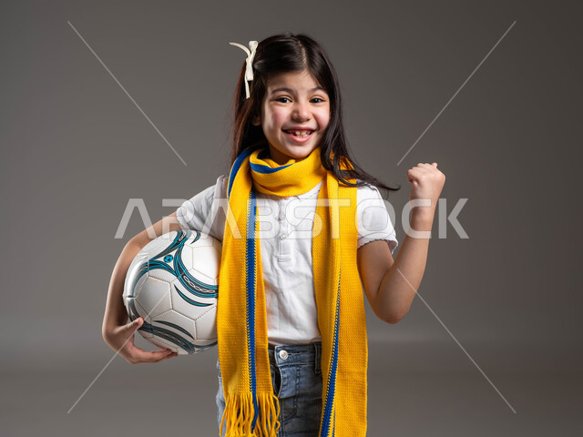 رفع اليد بكف مقبوض للأعلى بتعابير الانتصار، تشجيع الفريق السعودي لكرة القدم ، أنشطة رياضية ترفيهية للأطفال, بورتريه لفتاة عربية خليجية سعودية ترتدي الوشاح الأصفر وتحمل بيدها كرة قدم, النظر إلى الكاميرا بإيماءات تدل على الحماس والقوة، خلفية رمادية