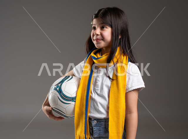 إيماءات الاندماج و الاعجاب, بورتريه لفتاة عربية خليجية سعودية مبتسمة ترتدي الوشاح الأصفر وتحمل بيدها كرة القدم، متابعة وتشجيع الفريق المفضل بتعابير الرضا و السرور، أنشطة رياضية ترفيهية للأطفال, خلفية رمادية