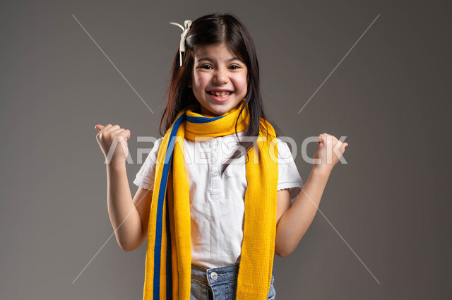 النظر إلى الكاميرا بإيماءات التشجيع و الحماس, بورتريه لفتاة خليجية عربية سعودية مبتسمة ترتدي الوشاح الأصفر و ترفع يديها إلى الجانب بكفين مقبوضتين بتعابير الحماس لفوز الفريق, أنشطة رياضية ترفيهية للأطفال، خلفية رمادية