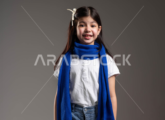 النظر إلى الكاميرا بإيماءات السرور، بورتريه لفتاة عربية سعودية خليجية مبتسمة ترتدي الوشاح الأزرق و تقف باستقامة بتعابير الرضا و الشعور بالسعادة، مفهوم الاهتمام بالمظهر الخارجي، خلفية رمادية