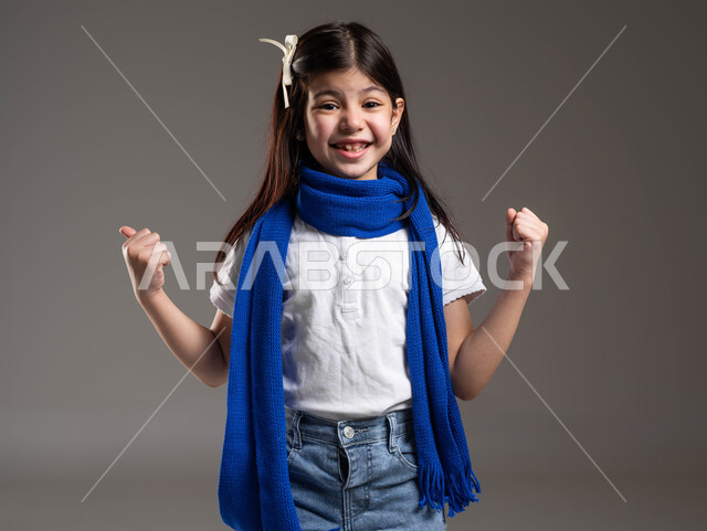 النظر إلى الكاميرا بإيماءات الحماس و التشجيع, بورتريه لفتاة خليجية عربية سعودية ترتدي الوشاح الأزرق ترفع يديها للأعلى بكفين مقبوضتين بتعابير النصر و الفوز, خلفية رمادية