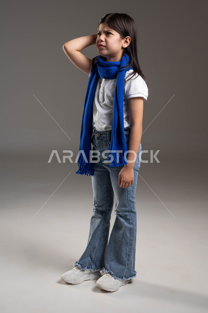 محاولة تذكر شيء معين, النظر إلى الأعلى بإيماءات الحيرة والحزن, بورتريه مقرب لفتاة عربية خليجية سعودية ترتدي الوشاح الأزرق وتضع يدها على رأسها بتعابير القلق و التفكير, الجسم بالطول الكامل, خلفية رمادية